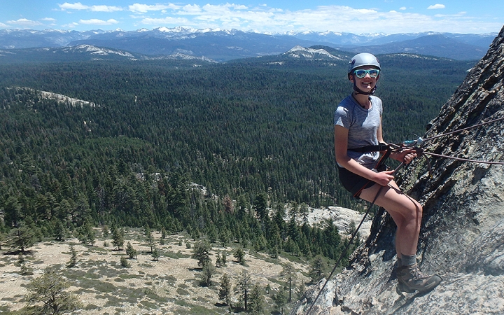 A young person wearing safety gear pauses to smile at the camera while rappelling a rock wall. There is a vast wooded and mountainous landscape in the background.